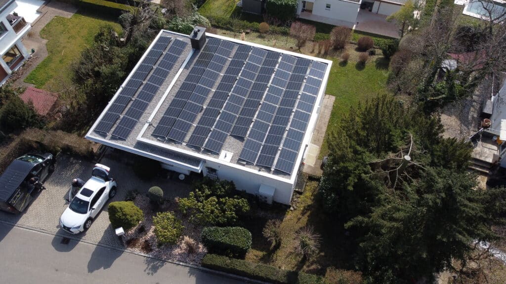 Dach mit Solarpanelen auf einem Wohnhaus, umgeben von Gartenanlagen und parkenden Autos. Die Sonnenkollektoren sind gut sichtbar und tragen zur nachhaltigen Energieerzeugung bei.